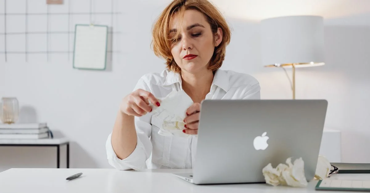 Femme portant une chemise blanche assise à un bureau avec un ordinateur portable, semblant frustrée avec des papiers froissés.