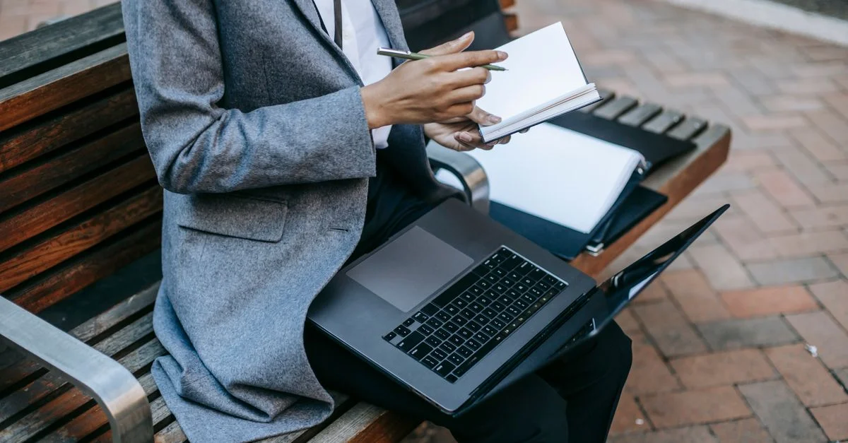 Femme d'affaires prenant des notes et utilisant un ordinateur portable sur un banc en plein air.