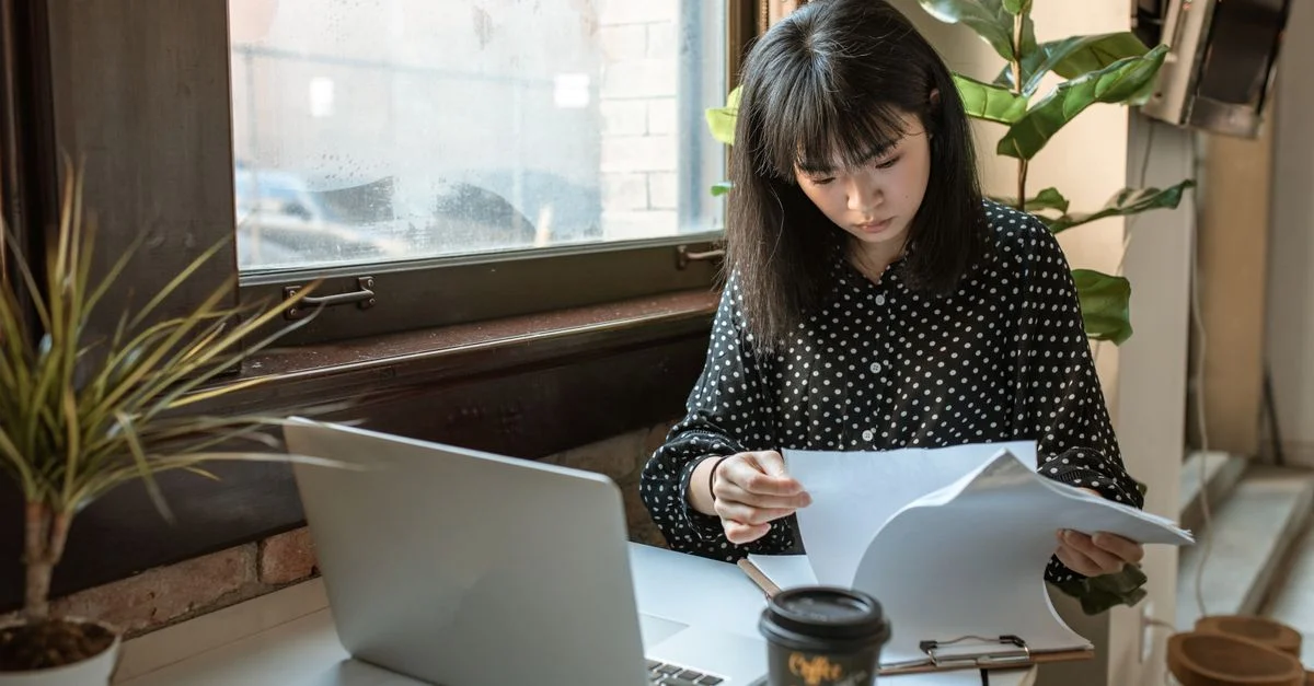 Femme d'affaires asiatique examinant des documents dans un bureau moderne avec lumière naturelle.